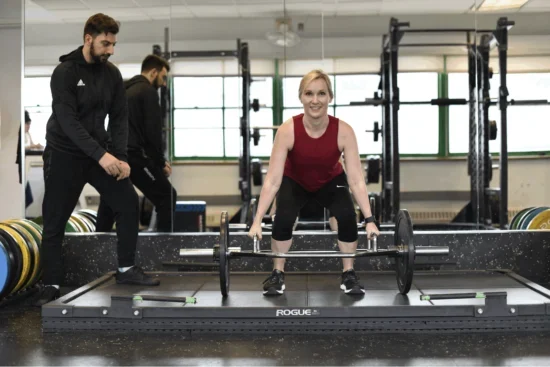A woman in athletic attire performs a barbell deadlift in a gym while an Asphalt Green trainer provides guidance on form.