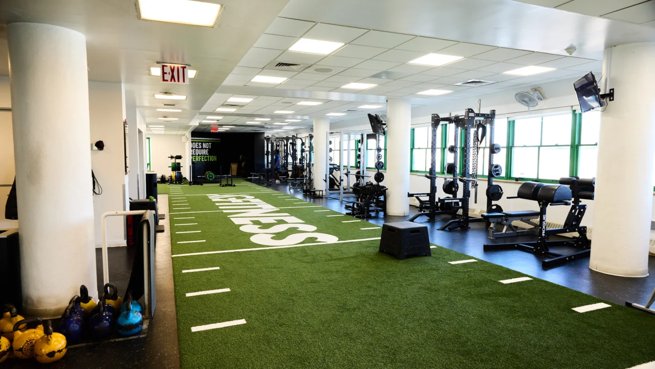 Spacious gym on the Upper East Side featuring turf flooring, white columns, weightlifting equipment, kettlebells, and an emergency exit sign on the ceiling.