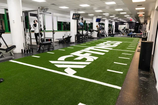 A gym interior with a turf area labeled "Fitness," weightlifting equipment, and exercise machines. People are working out in the background.