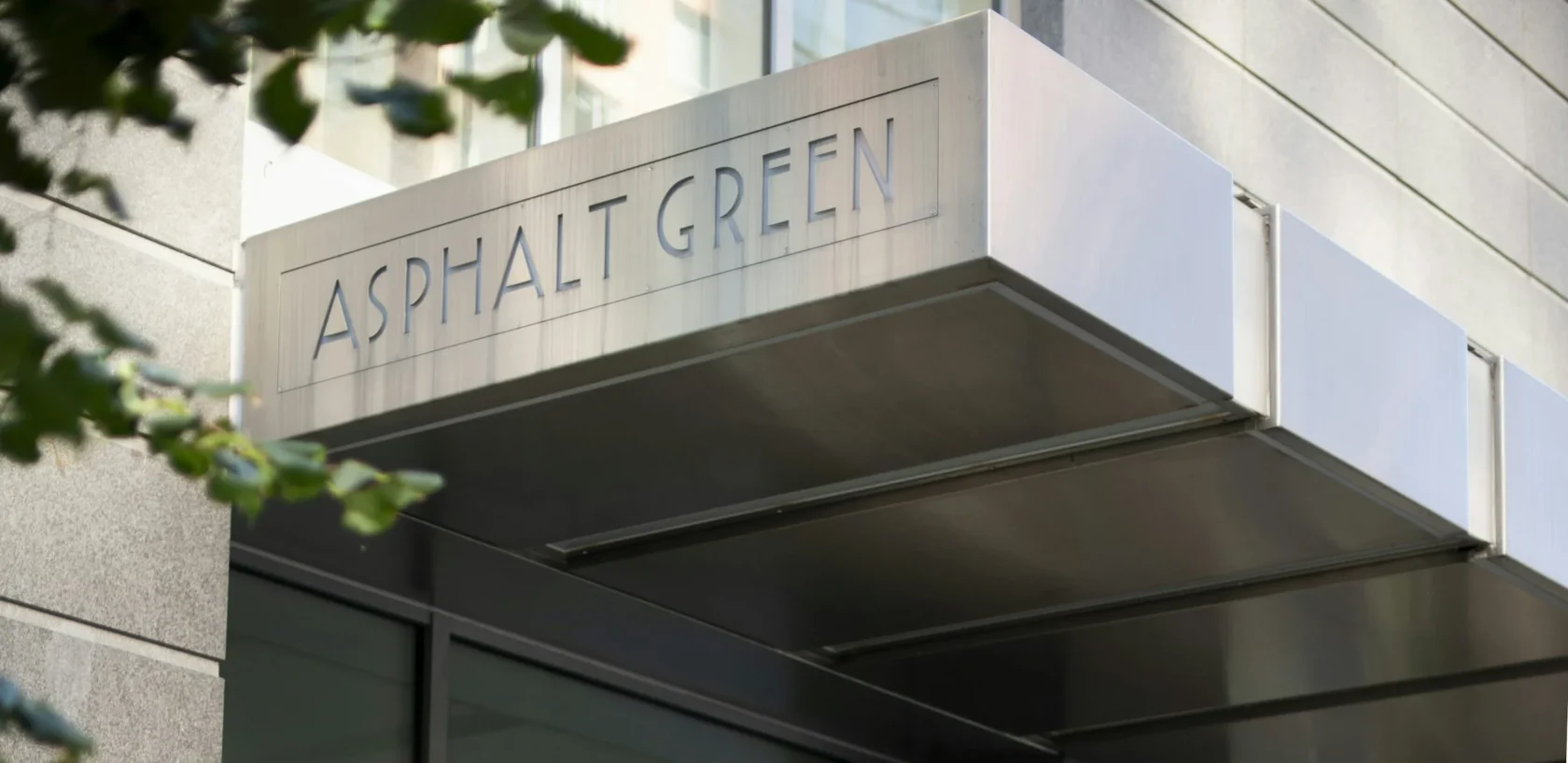 Sign reading "Asphalt Green" on a metal awning attached to a modern glass and stone building, with leaves slightly visible in the foreground.