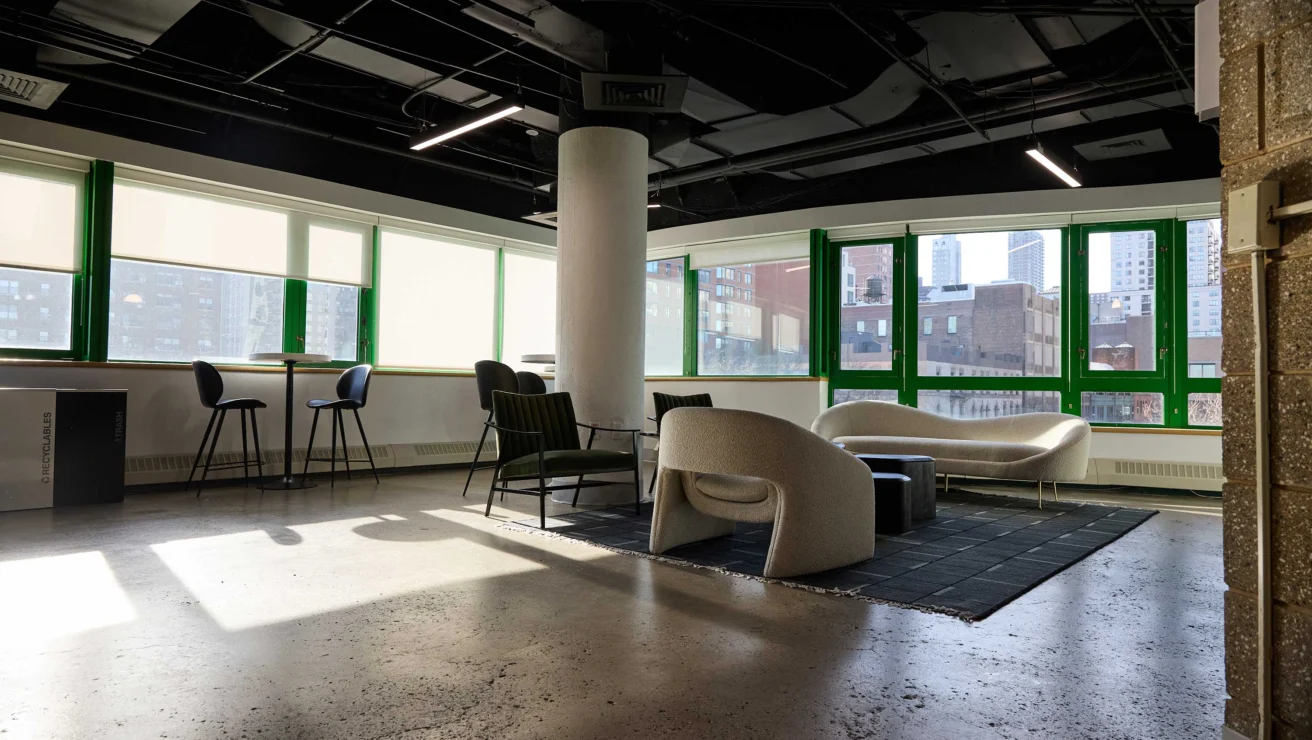 Modern office lounge on the Upper East Side with large windows, green frames, assorted seating, a column, and city view. Natural light fills the space, highlighting a polished concrete floor.
