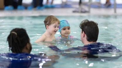 Two adults in a pool interact with a young boy and a girl wearing a swim cap. Other people are visible in the background near the water's edge.
