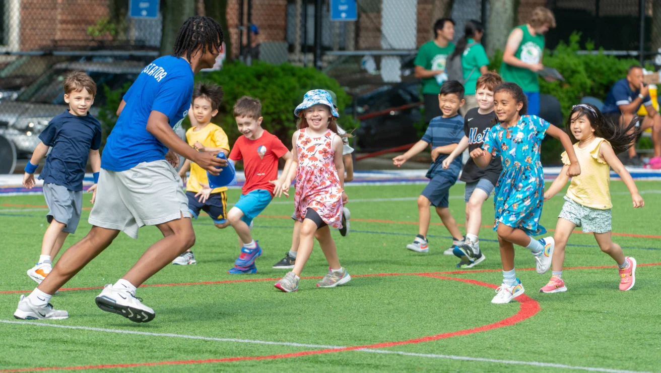 A group of children play a game with an adult on a grassy field. The adult holds a ball while the children run and smile.