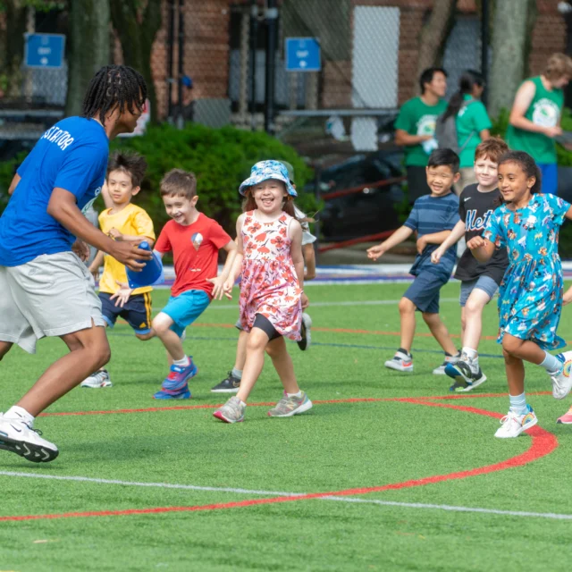 A group of children play a game with an adult on a grassy field. The adult holds a ball while the children run and smile.