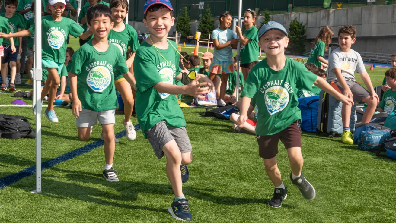 Children in green shirts running on a grassy field, smiling, with a group sitting in the background.
