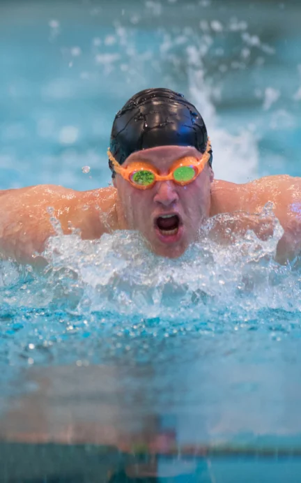 A swimmer in orange goggles and a black cap performs the butterfly stroke in a pool, splashing water around them.