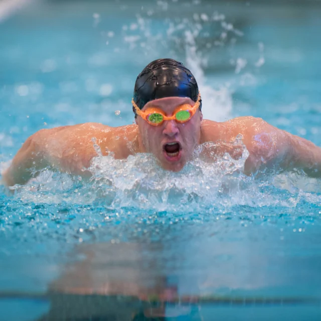 A swimmer in orange goggles and a black cap performs the butterfly stroke in a pool, splashing water around them.