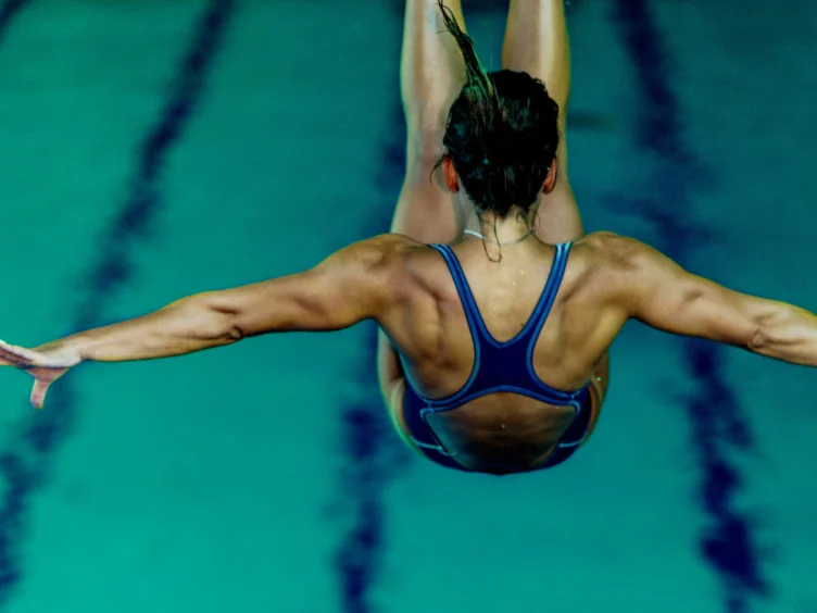 A diver in a blue swimsuit is captured mid-dive, arms outstretched over a swimming pool.