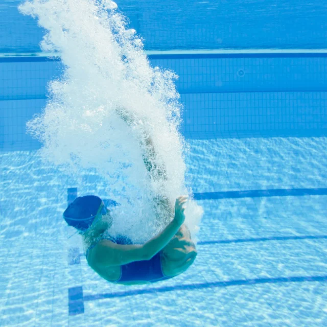 A swimmer in a blue swimsuit and goggles is submerged underwater, creating a large splash.