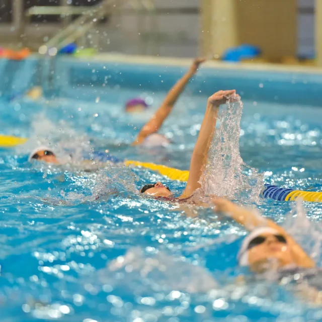 Swimmers in a pool practice synchronized backstroke, creating splashes as they move in unison.