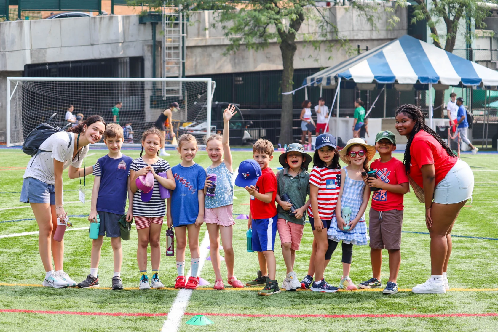 A group of children and adults pose together on a sports field with soccer goals in the background. Some children are wearing hats, and a tent is visible in the distance.