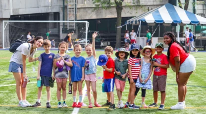 A group of children and adults pose together on a sports field with soccer goals in the background. Some children are wearing hats, and a tent is visible in the distance.
