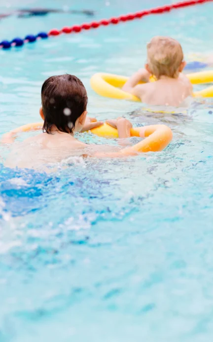 Children in a swimming pool use yellow pool noodles for support, with lanes marked by floating ropes.