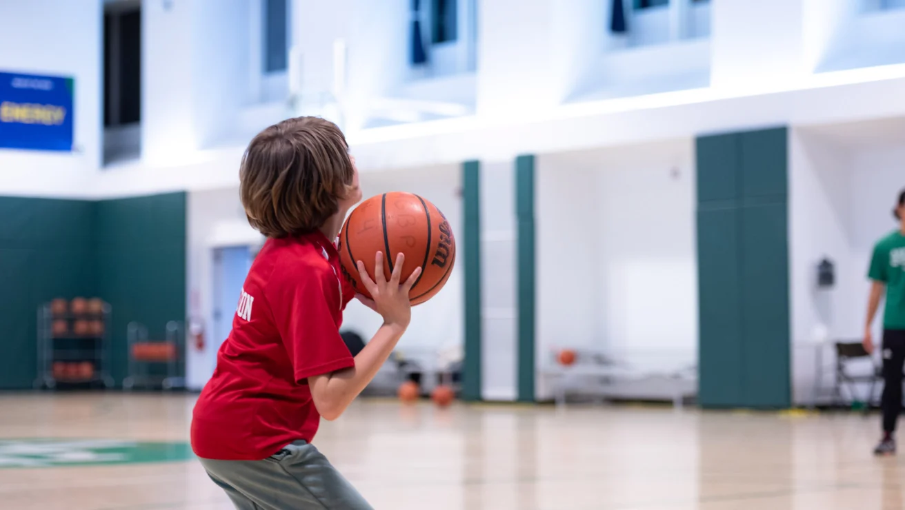 A child in a red shirt prepares to shoot a basketball in an indoor gym.