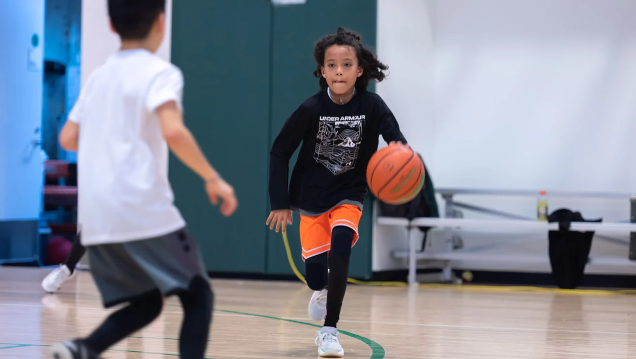 A child in a black shirt dribbles a basketball on an indoor court, approaching another child in a white shirt.
