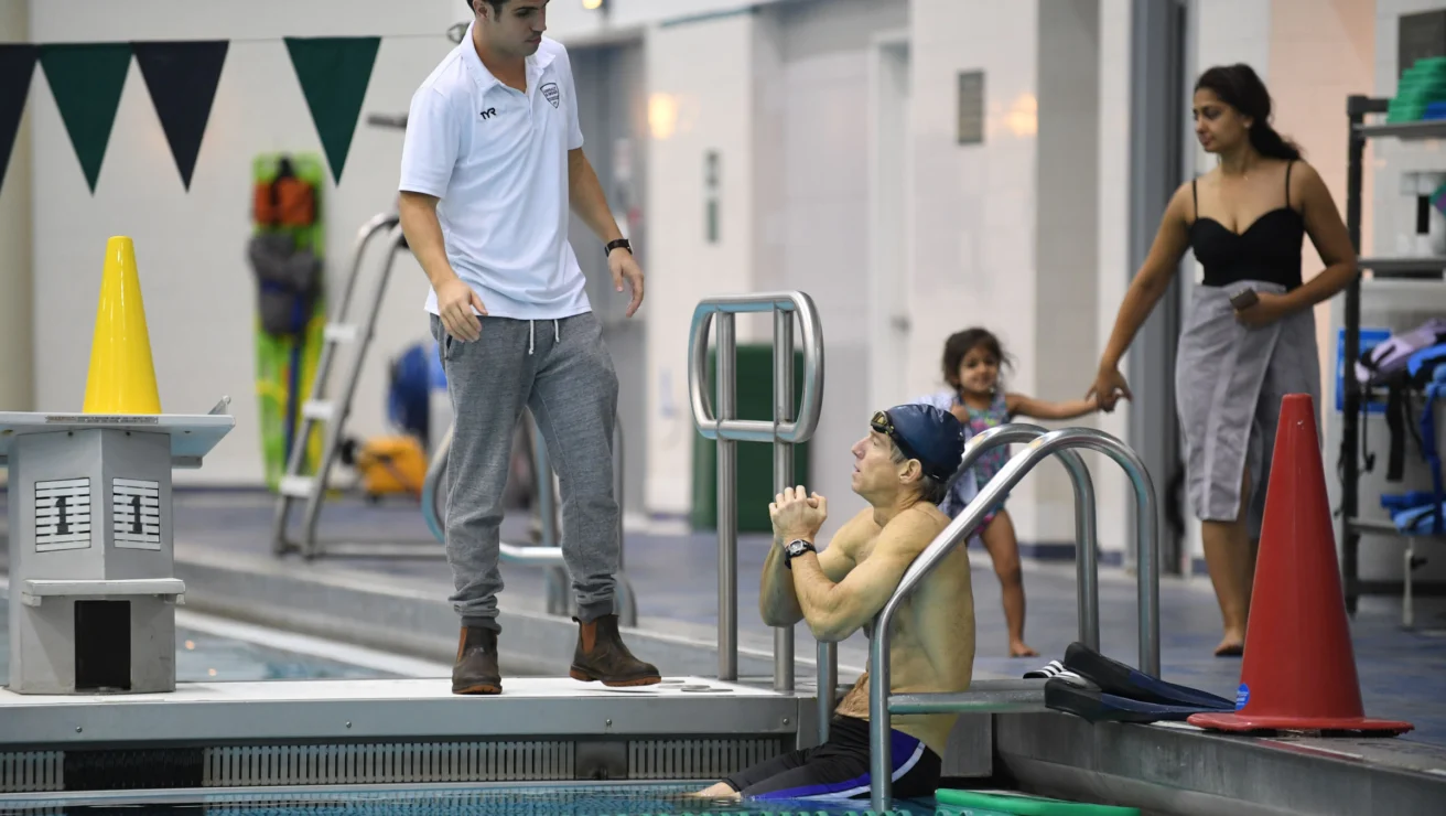 A swimmer wearing goggles sits at the edge of a pool, listening to a person standing nearby. A woman and a child walk in the background.