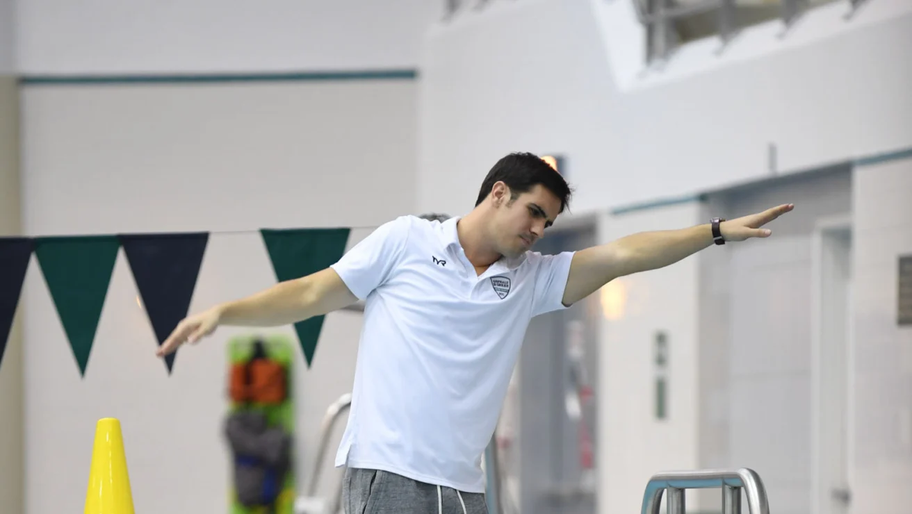 A coach instructs a swimmer at the edge of an indoor pool. The swimmer is sitting and listening attentively while the coach gestures with an outstretched arm.