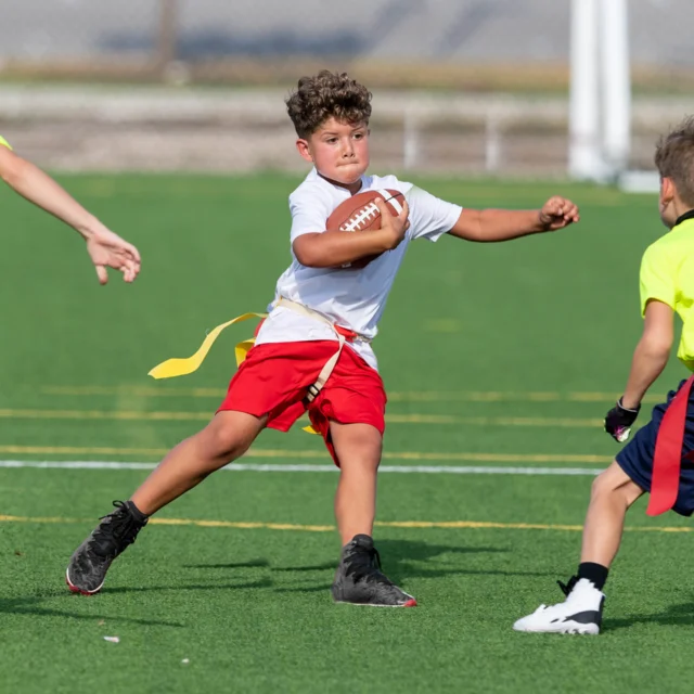 A child wearing a white shirt and red shorts plays flag football, holding the ball while evading two opponents on a field.