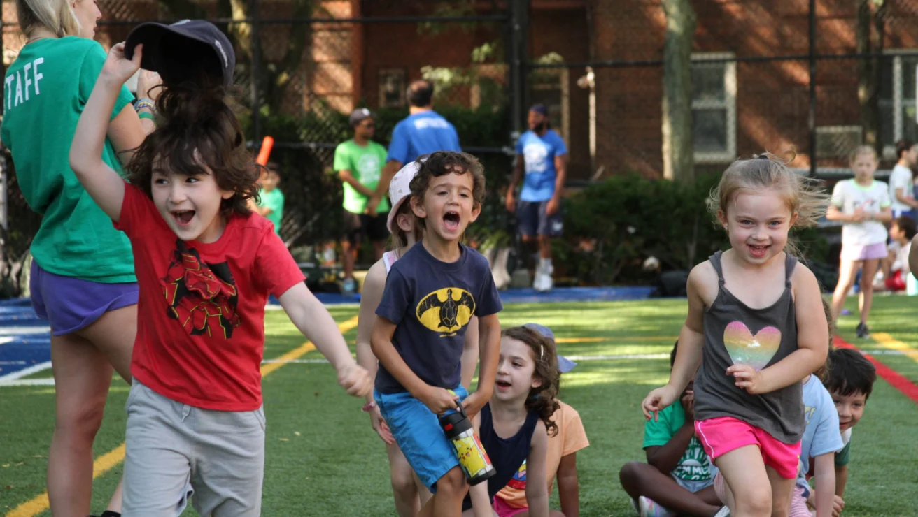 Children happily running and playing on a grass field during a sunny day, with a few adults in the background watching.