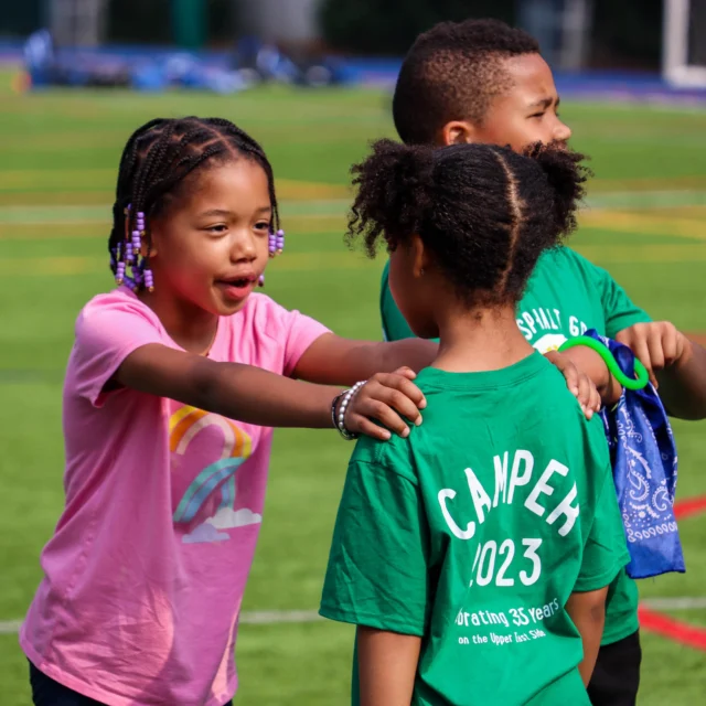 Children play a game on a sports field. One girl in a pink shirt talks to another girl in a green shirt with "CAMPS 2023" on the back. Other children stand nearby.