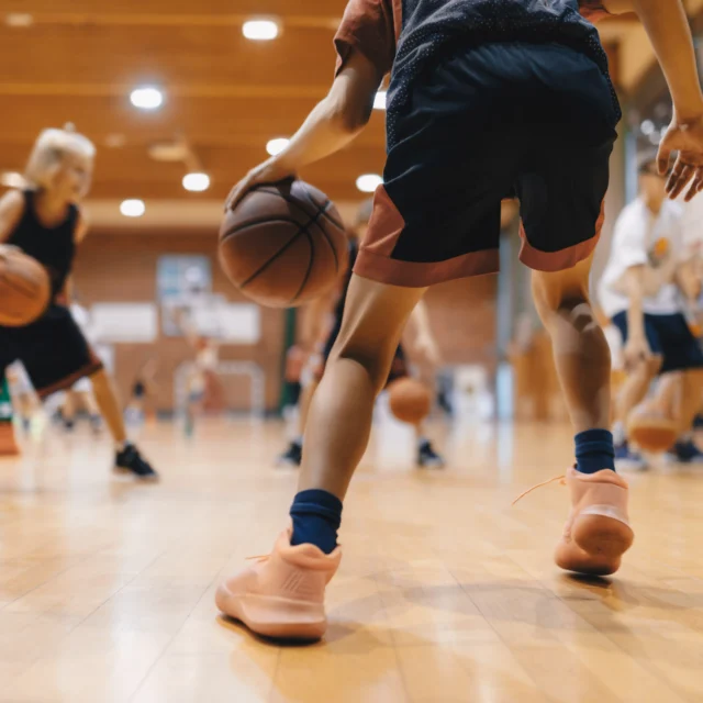 A group of people dribbling basketballs in a gym, focusing on their legs and sneakers on the wooden floor.
