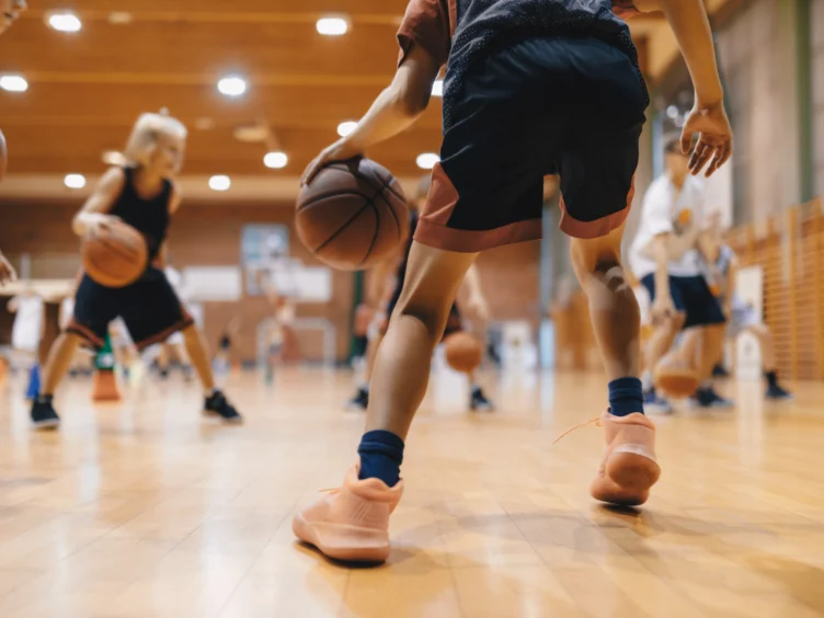 A group of people dribbling basketballs in a gym, focusing on their legs and sneakers on the wooden floor.