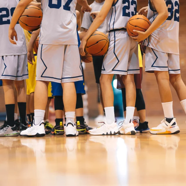 A group of basketball players in uniform huddle together on a gym court, holding basketballs.