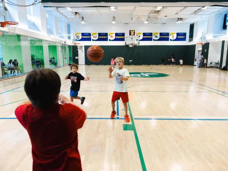 Children playing basketball in an indoor gym, with one child catching the ball in mid-air.