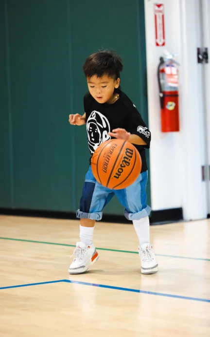 A young boy in a black shirt dribbles a basketball in a gym.