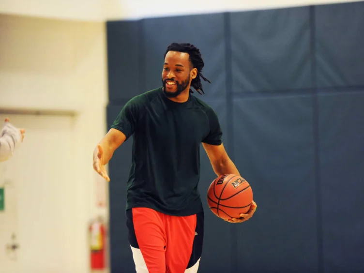 A person holding a basketball smiles and extends a hand on an indoor court.