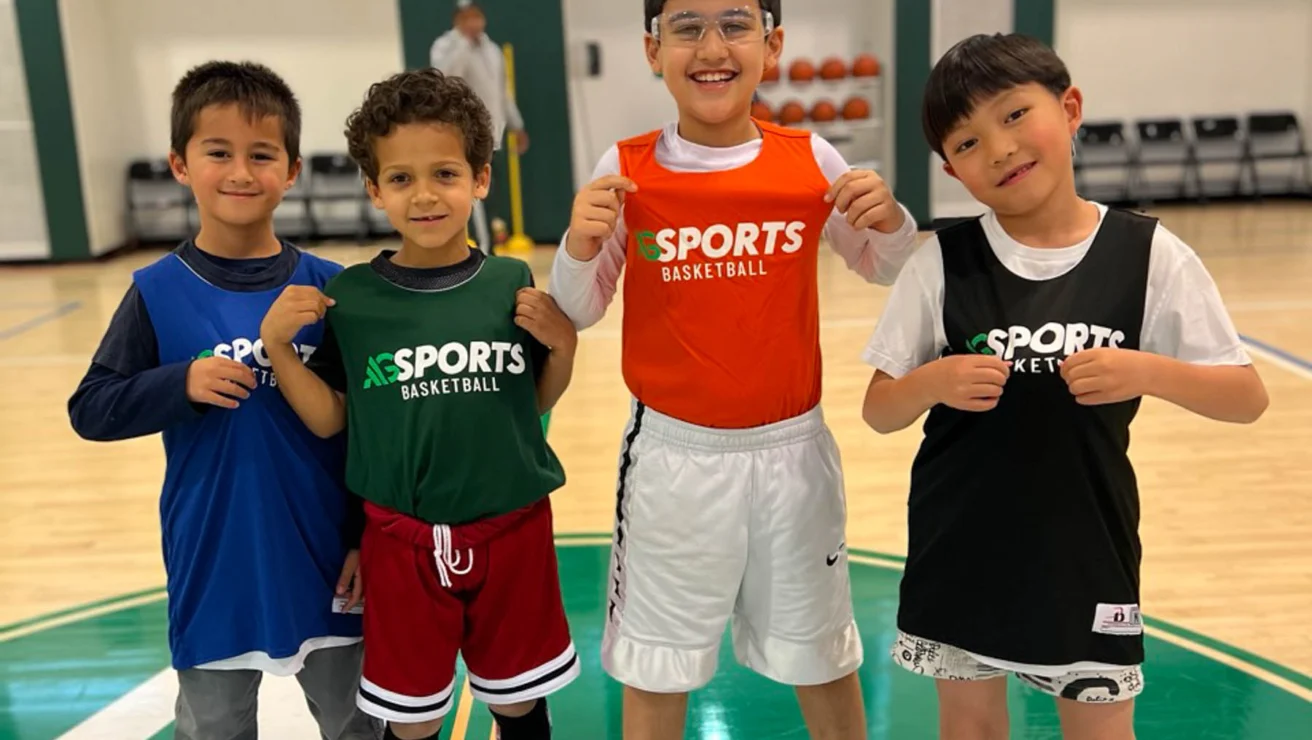 Four children in sports clothing, smiling and standing on a basketball court. They each wear a jersey with the word "Sports" on it.