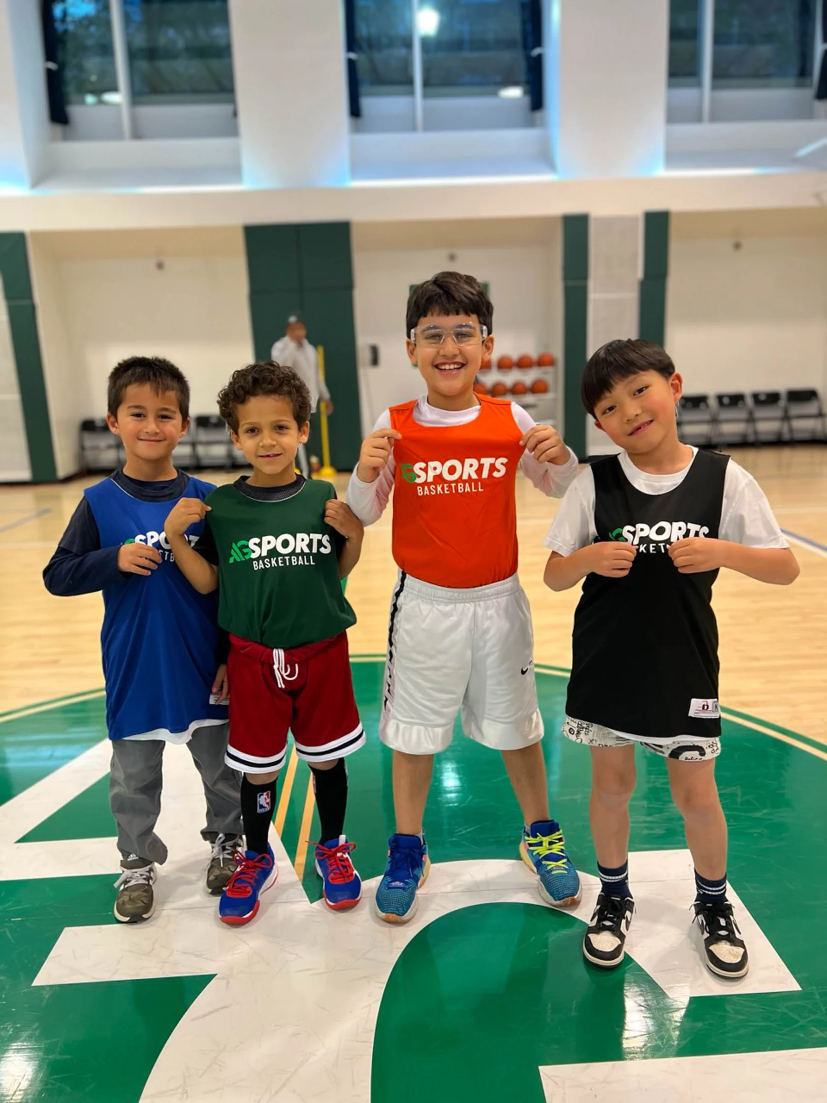 Four children in sports clothing, smiling and standing on a basketball court. They each wear a jersey with the word "Sports" on it.