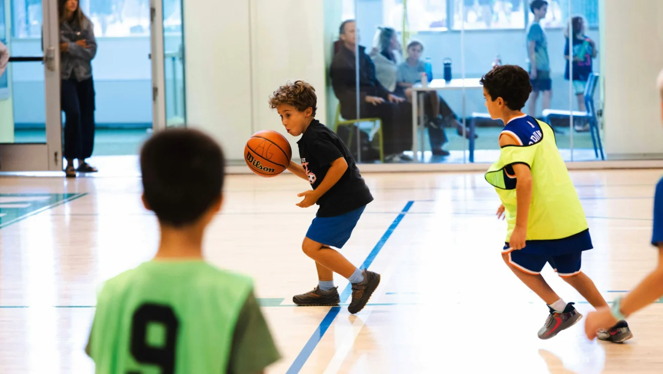 Young kids playing basketball indoors, with one child dribbling the ball while others guard him. Spectators can be seen in the background.