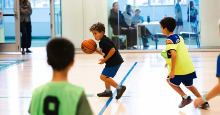Young kids playing basketball indoors, with one child dribbling the ball while others guard him. Spectators can be seen in the background.