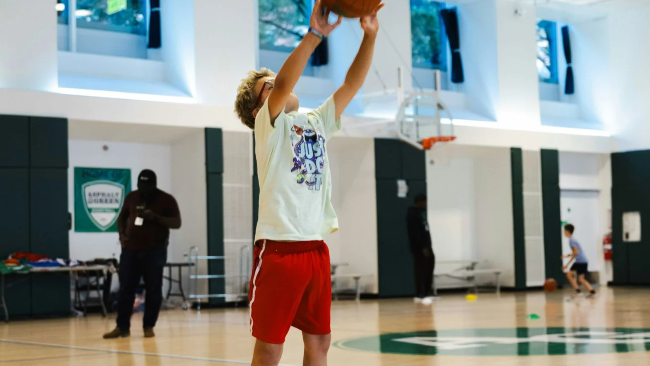A person in red shorts and orange sneakers jumps to shoot a basketball in an indoor court with others in the background.