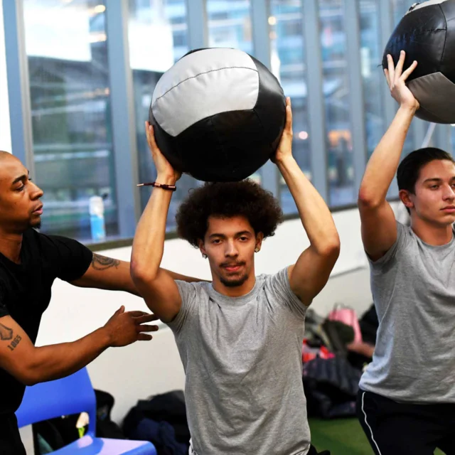 Three people exercise indoors; an instructor assists two individuals holding medicine balls overhead.