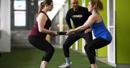 Two women squat while holding a weight plate together, guided by a fitness instructor in a gym setting.