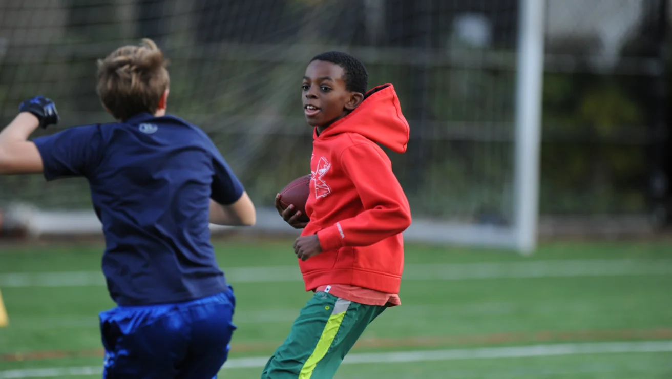Two boys playing football on a field, one in a red hoodie holding the ball, and the other in a blue shirt running nearby.