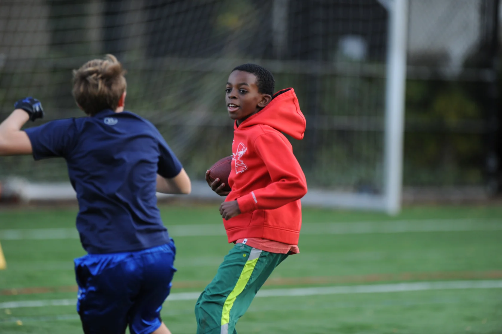 Two boys playing football on a field, one in a red hoodie holding the ball, and the other in a blue shirt running nearby.