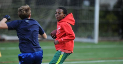 Two boys playing football on a field, one in a red hoodie holding the ball, and the other in a blue shirt running nearby.