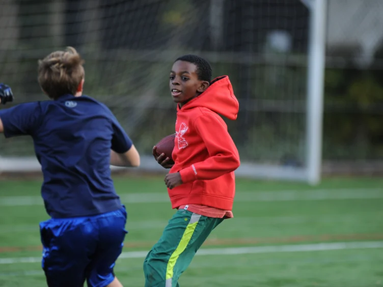 Two boys playing football on a field, one in a red hoodie holding the ball, and the other in a blue shirt running nearby.