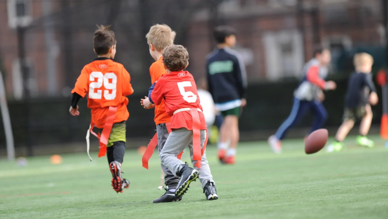 Kids playing flag football on a grassy field, two wearing orange jerseys with numbers 29 and 5. A football is in mid-air.