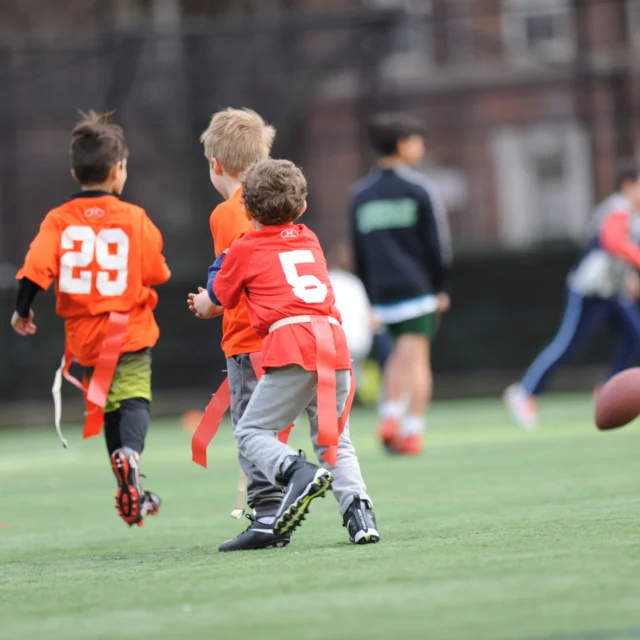 Kids playing flag football on a grassy field, two wearing orange jerseys with numbers 29 and 5. A football is in mid-air.