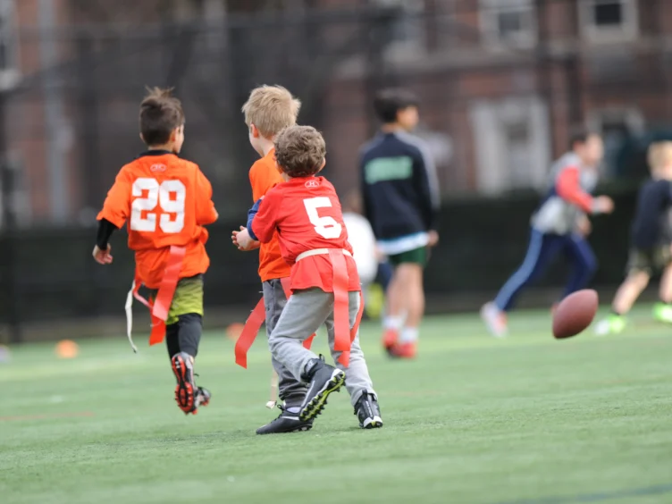Kids playing flag football on a grassy field, two wearing orange jerseys with numbers 29 and 5. A football is in mid-air.