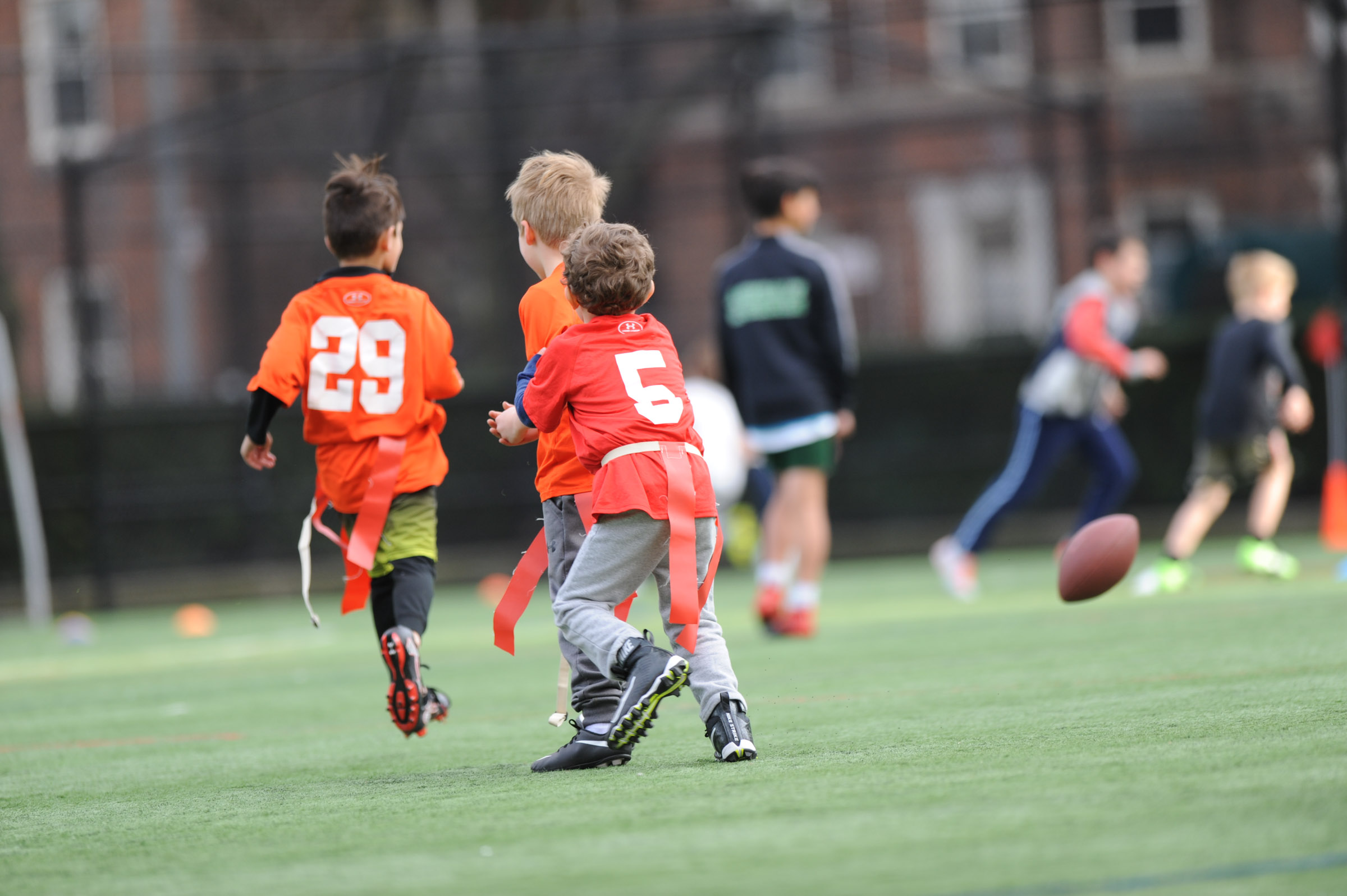 Kids playing flag football on a grassy field, two wearing orange jerseys with numbers 29 and 5. A football is in mid-air.