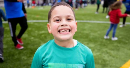 Child in a green shirt smiling at the camera on a grassy field with people in the background.