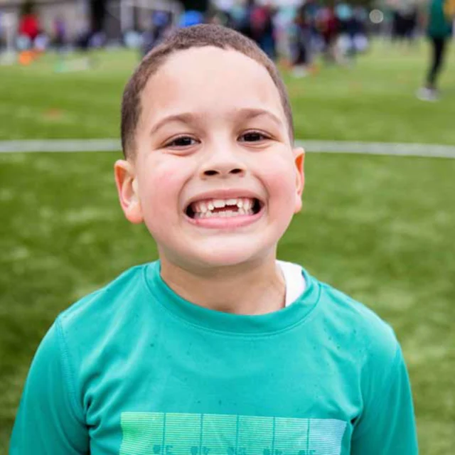 Child in a green shirt smiling at the camera on a grassy field with people in the background.