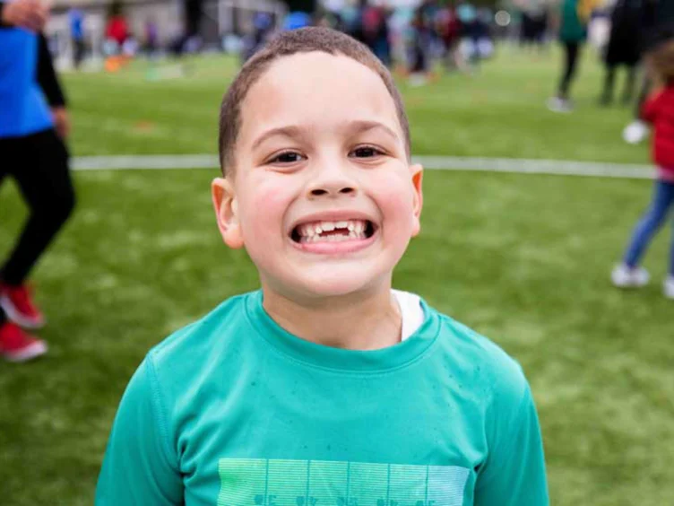 Child in a green shirt smiling at the camera on a grassy field with people in the background.