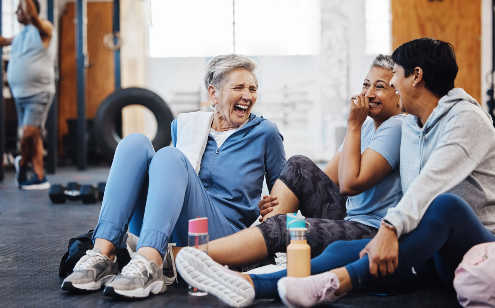 Three women in athletic wear sit on a gym floor, laughing and talking. Water bottles are beside them, and gym equipment is visible in the background.