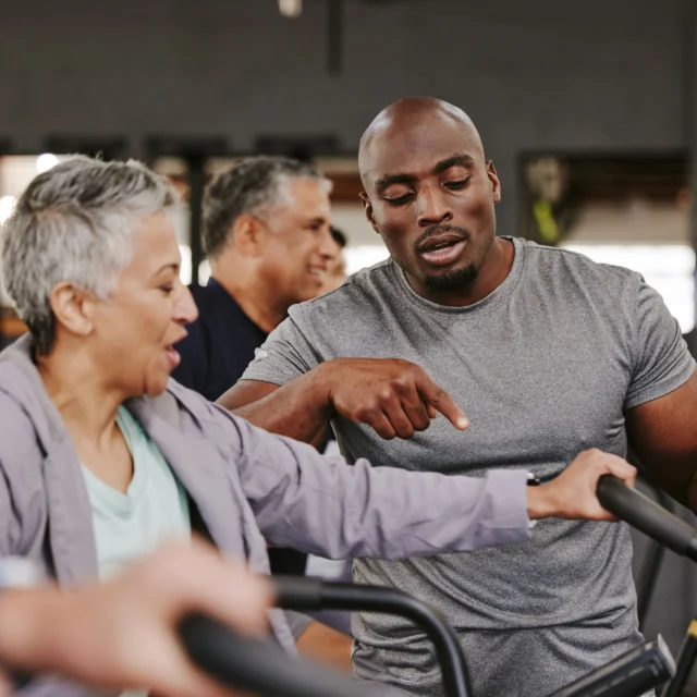 A man instructs an older woman on exercise equipment in a gym, with two other people in the background.